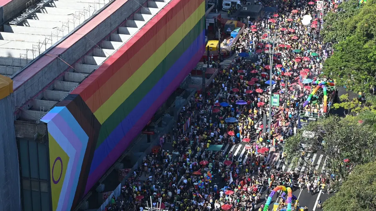 A massive crowd fills the street for what appears to be the biggest pride parade, with a giant rainbow flag draped over a building.