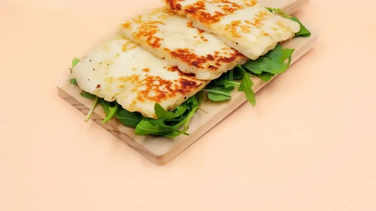 Trzy grillowane plastry sera halloumi, z czego jest, ułożone na desce z rukolą.