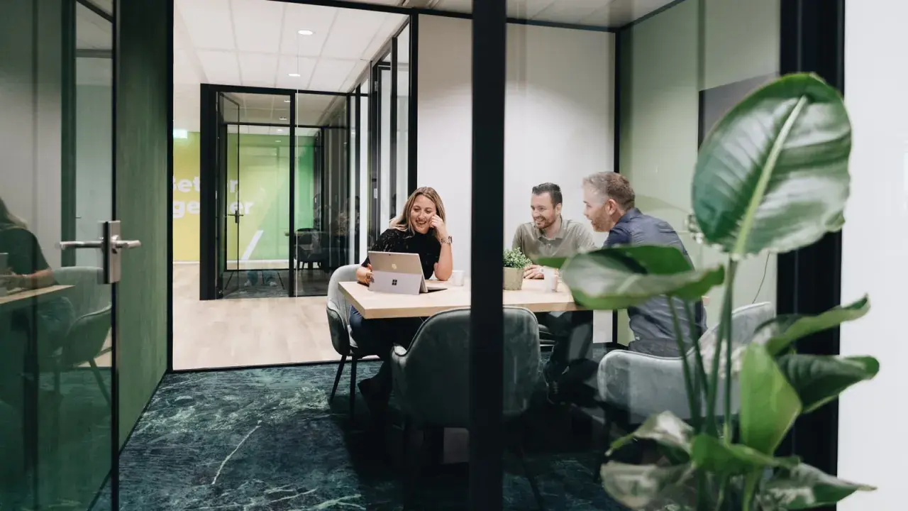 Tre kollegor skrattar vid ett mötesbord i en modern, inredd verkstad. En kvinna visar något på sin laptop.