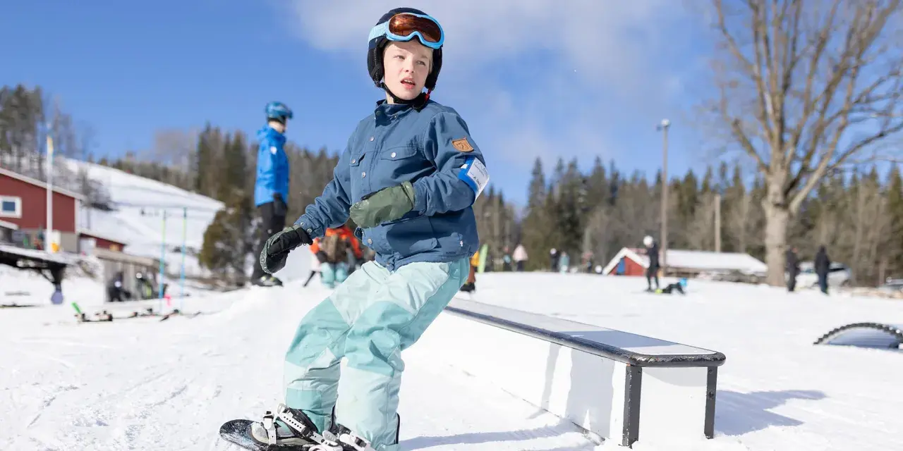 Ungdom åker snowboard i backen, redo för nästa trick. Solen skiner och snön ligger vit.