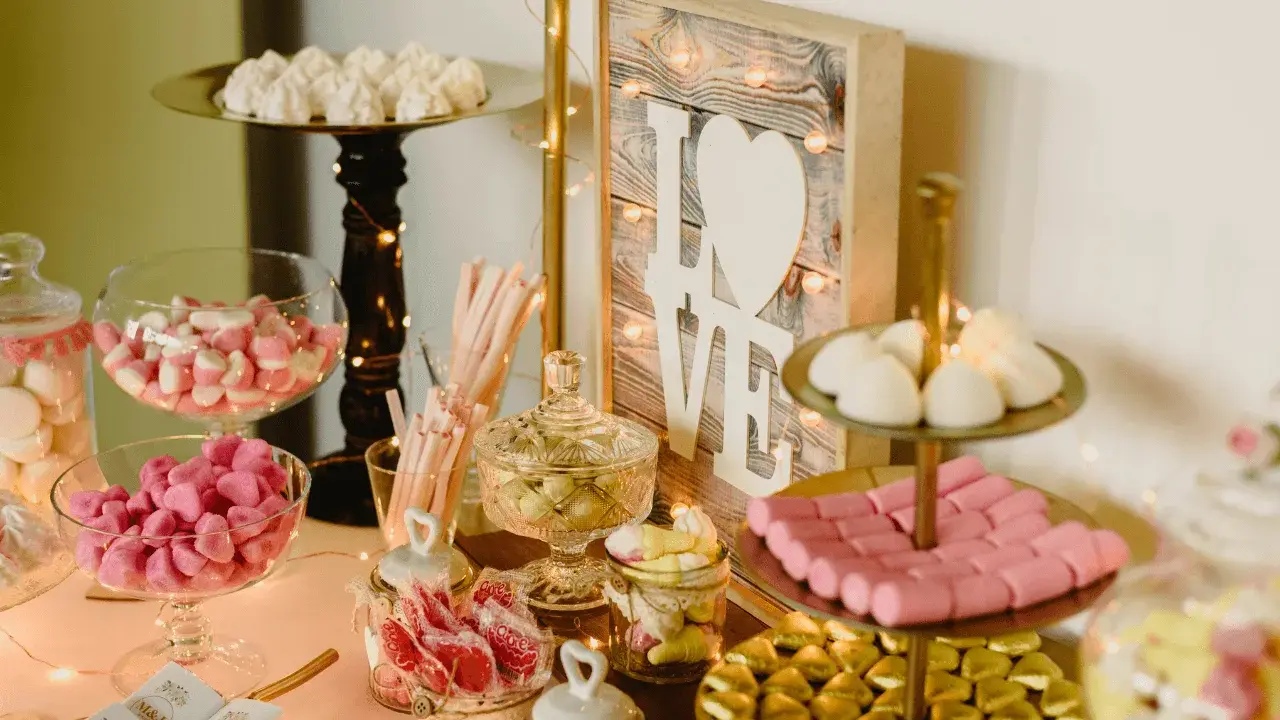 Un candy bar lleno de dulces rosas y blancos, con un letrero de "LOVE" y luces cálidas.