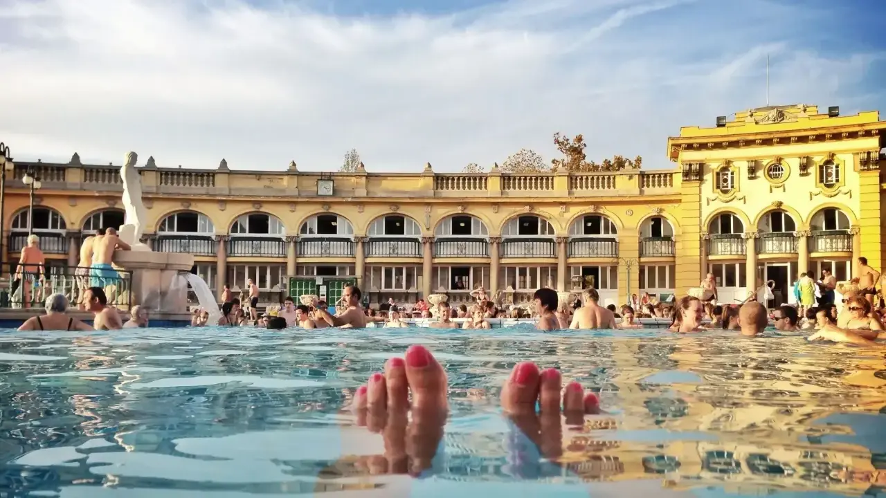 Fötter sticker upp ur vattnet i ett soligt budapest spa. Människor badar och njuter av den vackra arkitekturen.