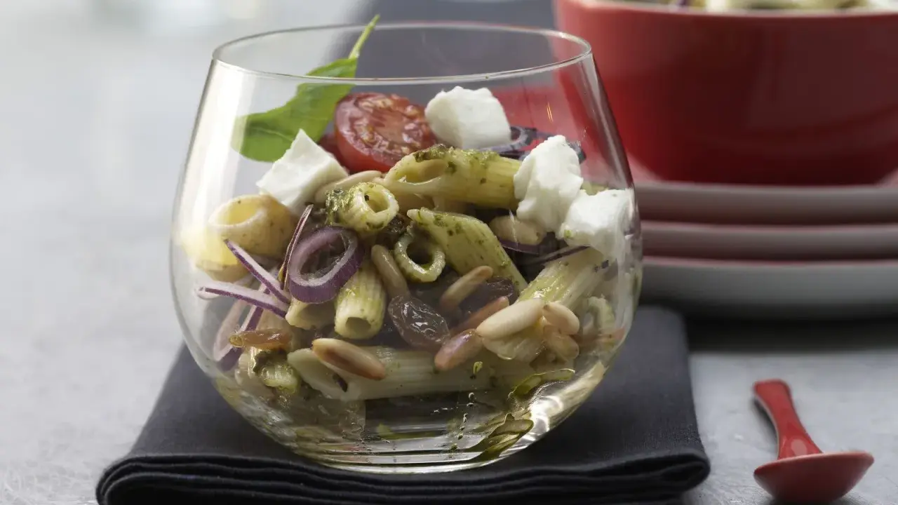 Ein köstlicher Nudel Pesto Salat mit Mozzarella, Pinienkernen, Rosinen und roten Zwiebeln in einem Glas serviert.