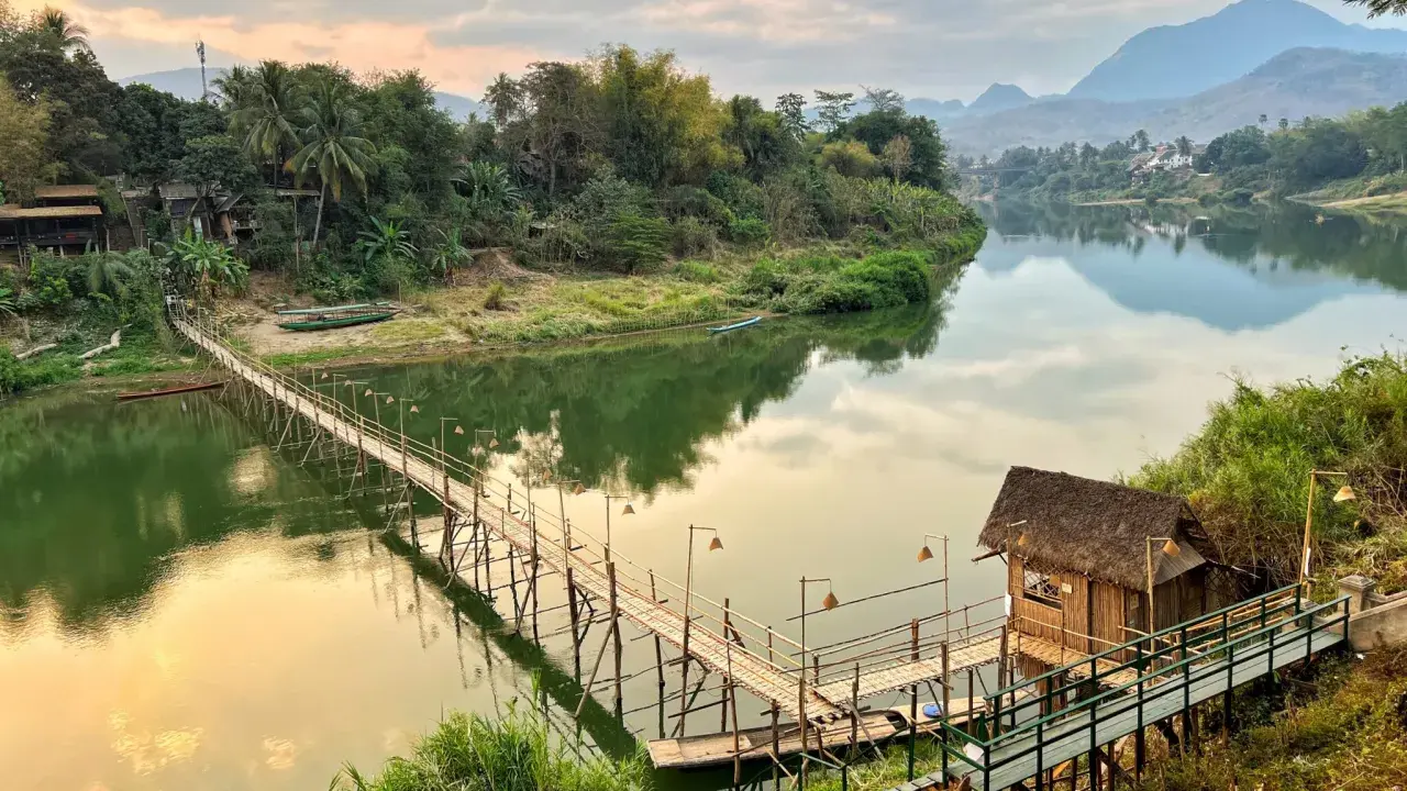 Drewniany mostek nad rzeką, bambusowa chatka i góry w tle. To jedne z wielu **laos atrakcje** warte zobaczenia.