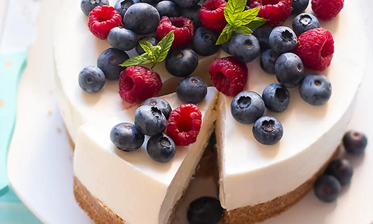 Cheesecake allo yogurt fresca e cremosa, decorata con lamponi e mirtilli succosi. Una fetta è già stata tagliata, invitando a gustarla.