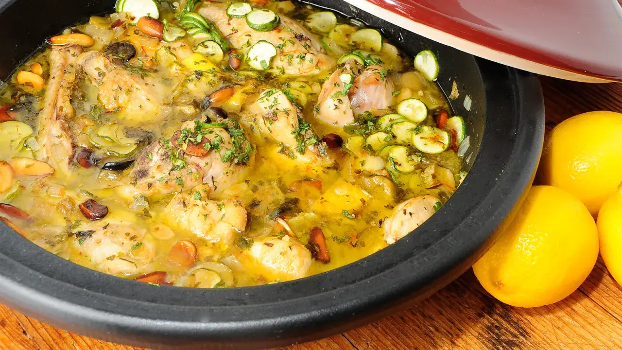 Tajín de pollo con calabacín, aceitunas y almendras, servido en una cazuela de barro, con limones frescos al lado.