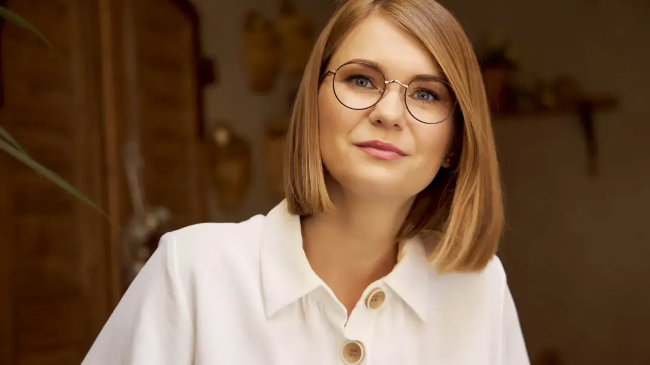 Kobieta w białej koszuli z guzikami, nosząca okulary w cienkich oprawkach, patrzy wprost.