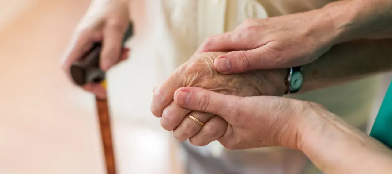 Dwie dłonie, jedna starsza, druga młodsza, trzymają się nawzajem. Laska w tle sugeruje trudności z poruszaniem się, być może związane z chorobą Parkinsona i jej dziedziczeniem.