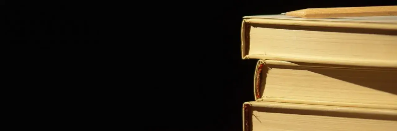Libri impilati, un libro in cima con una matita. La luce crea ombre, un po' come l'anastrofe figura retorica che inverte l'ordine per enfasi.
