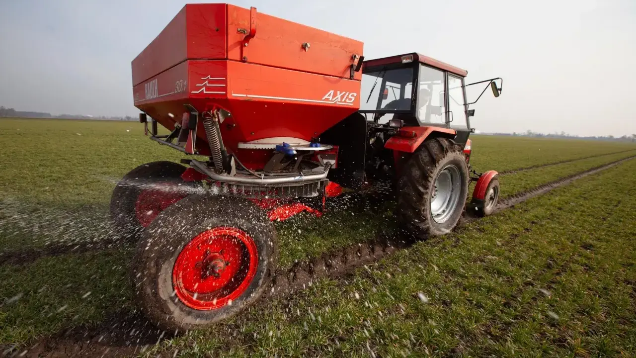 Czerwony traktor z rozsiewaczem nawozu na wiosennym polu. Odpowiedni nawóz pod warzywa na wiosnę to klucz do obfitych plonów.