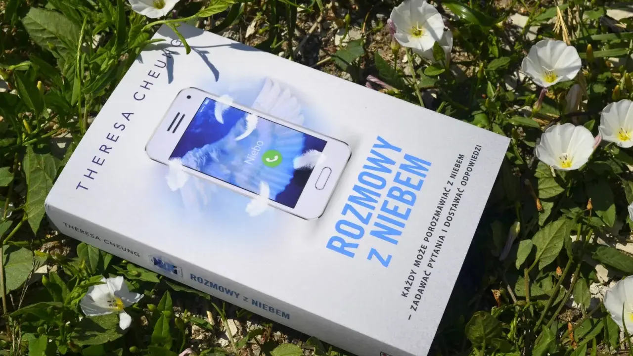 Książka "Rozmowy z niebem" leży wśród zieleni i białych kwiatów. Na okładce telefon z napisem "Niebo" i piórami.