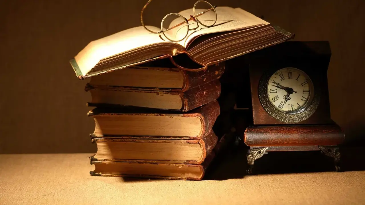 Un libro aperto con occhiali da lettura, una pila di antichi volumi e un orologio da tavolo. Un invito a leggere Catullo: "dammi mille baci".