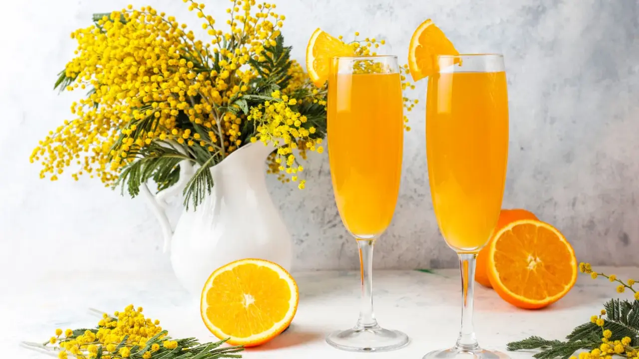 Dwa kieliszki orzeźwiającego drinka mimosa, ozdobione plasterkami pomarańczy, stoją obok bukietu żółtych kwiatów akacji.
