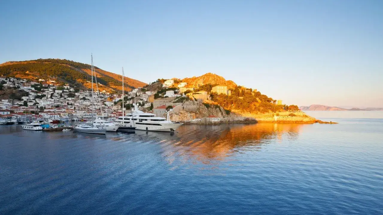 Solnedgång över hamnen på Hydra, Grekland. Lyxyachter guppar i det blå vattnet framför en vit stad som klättrar uppför en grönskande kulle.
