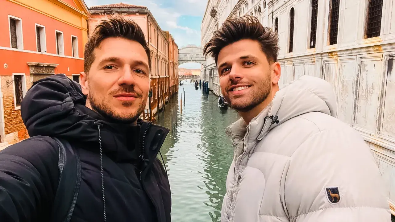 Two men enjoying a romantic gay Venice trip, smiling for a selfie with canals and historic buildings behind them.