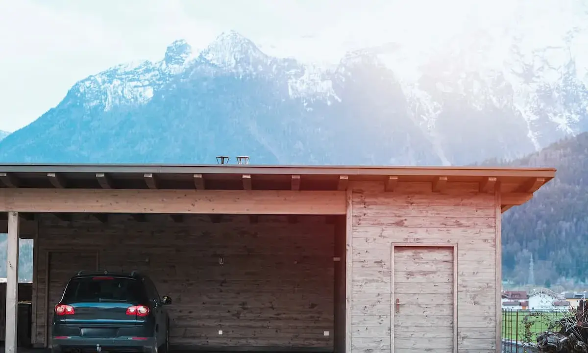En nybyggd carport i trä med en bil parkerad under. Bakom syns snöklädda berg.