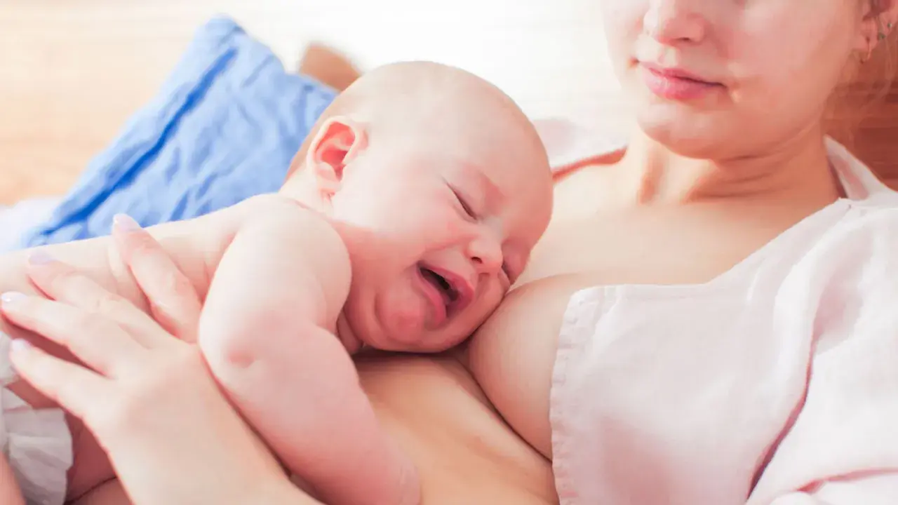 Ein Baby schreit beim Stillen, die Augen geschlossen, das Gesicht verzogen. Die Mutter hält es sanft.