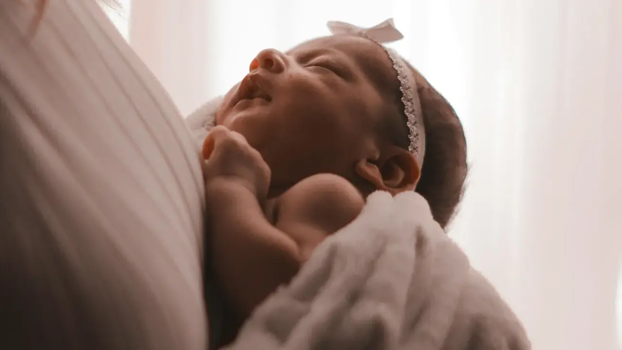 Un neonato avvolto in una coperta bianca, con un fiocco rosa in testa. I suoi piccoli tremori sono quasi impercettibili mentre dorme serenamente.