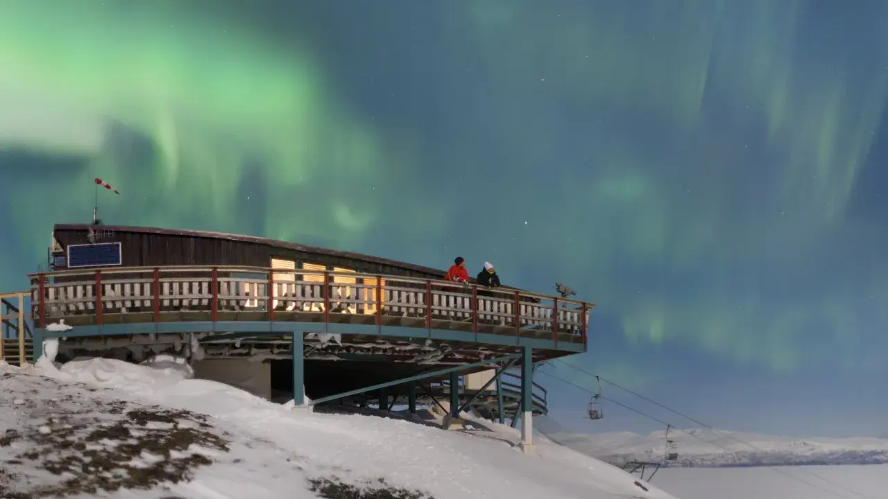 Två personer njuter av norrskenet från en aurora sky station i snöig fjällmiljö.