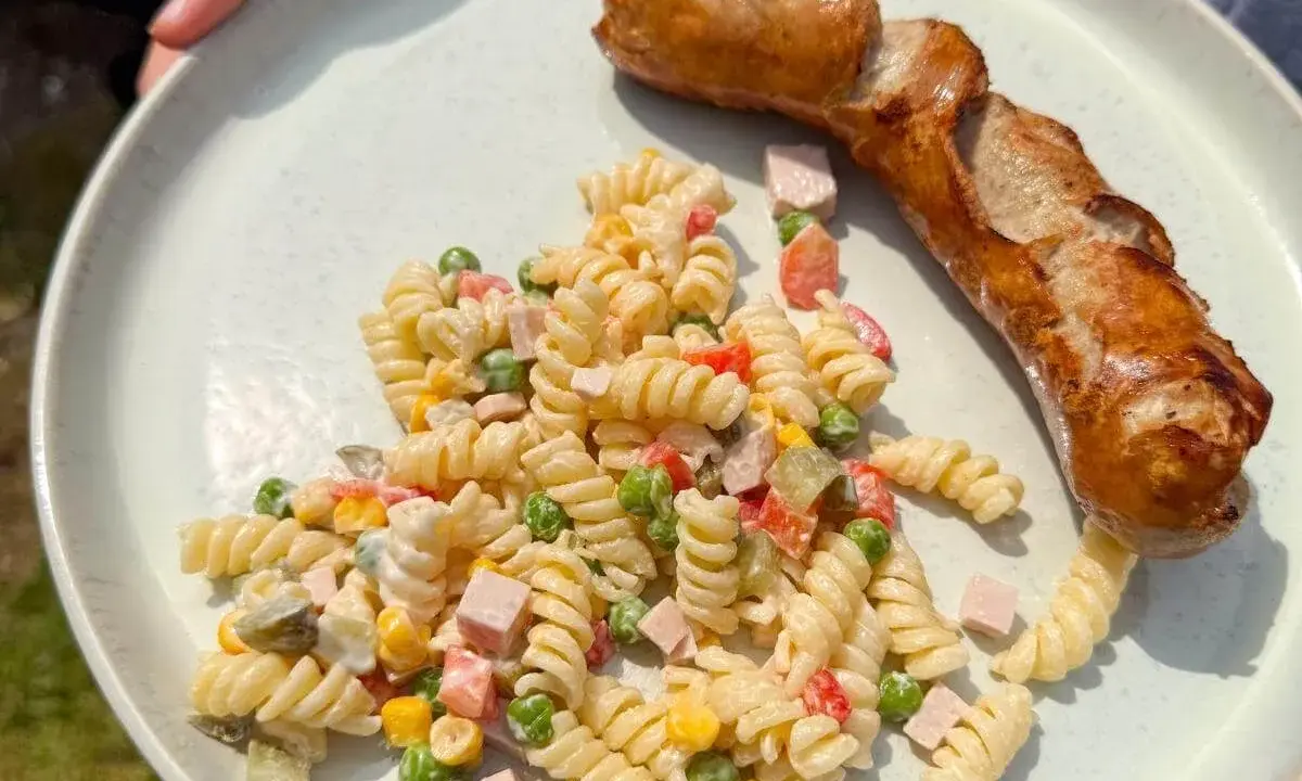 Gegrillte Wurst mit einem cremigen Nudelsalat als Beilage, garniert mit Erbsen, Mais und gewürfeltem Schinken.