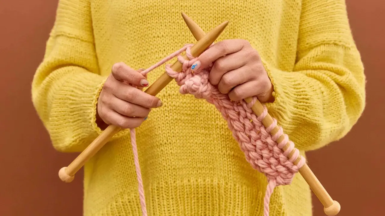 Manos tejiendo un suéter amarillo con hilo rosa, mostrando cómo hacer ochos de 4 puntos.