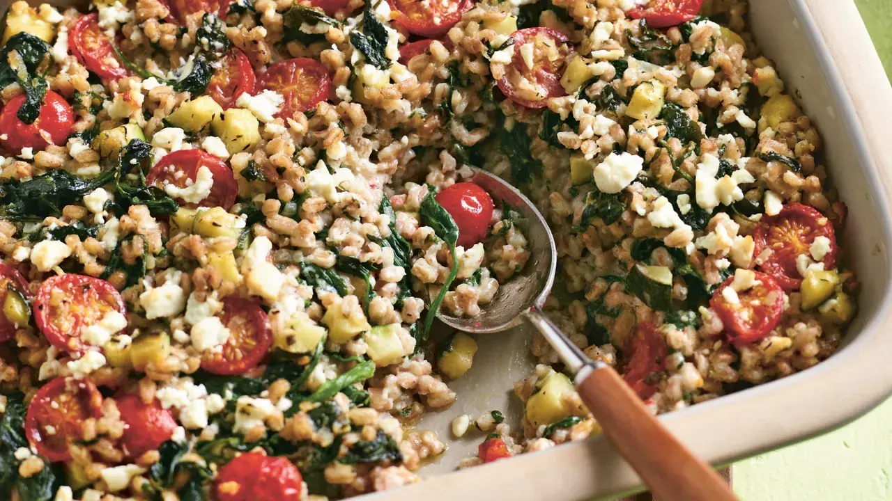 Herzhaftes Gericht mit gekochtem Dinkel, Tomaten, Zucchini und Feta. Ein Löffel hebt die köstliche Mischung aus der Auflaufform.