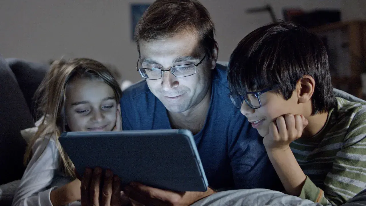 Padre y dos hijos ven una tablet, sonriendo. Podrían estar aprendiendo sobre los riesgos de las redes sociales o disfrutando de contenido familiar.