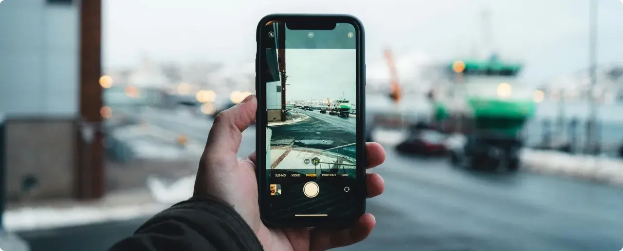 Dłoń trzyma aparat na telefon, rejestrując zimowy widok portu z zielonym statkiem i samochodami na drodze.