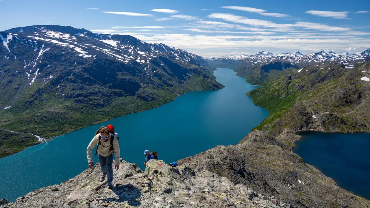 Vandrare klättrar på en bergskam med en turkos sjö nedanför. Besseggen svårighetsgrad är utmanande men belönande.