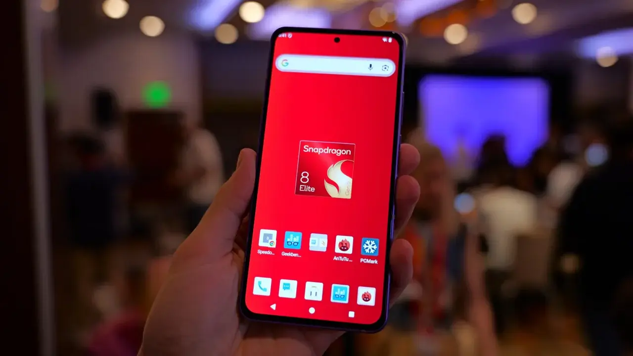 Dłoń trzyma smartfon z czerwonym tłem i ikoną Snapdragon 8 Elite. Widoczne ikony benchmarków, sugerujące testowanie wydajności procesora.