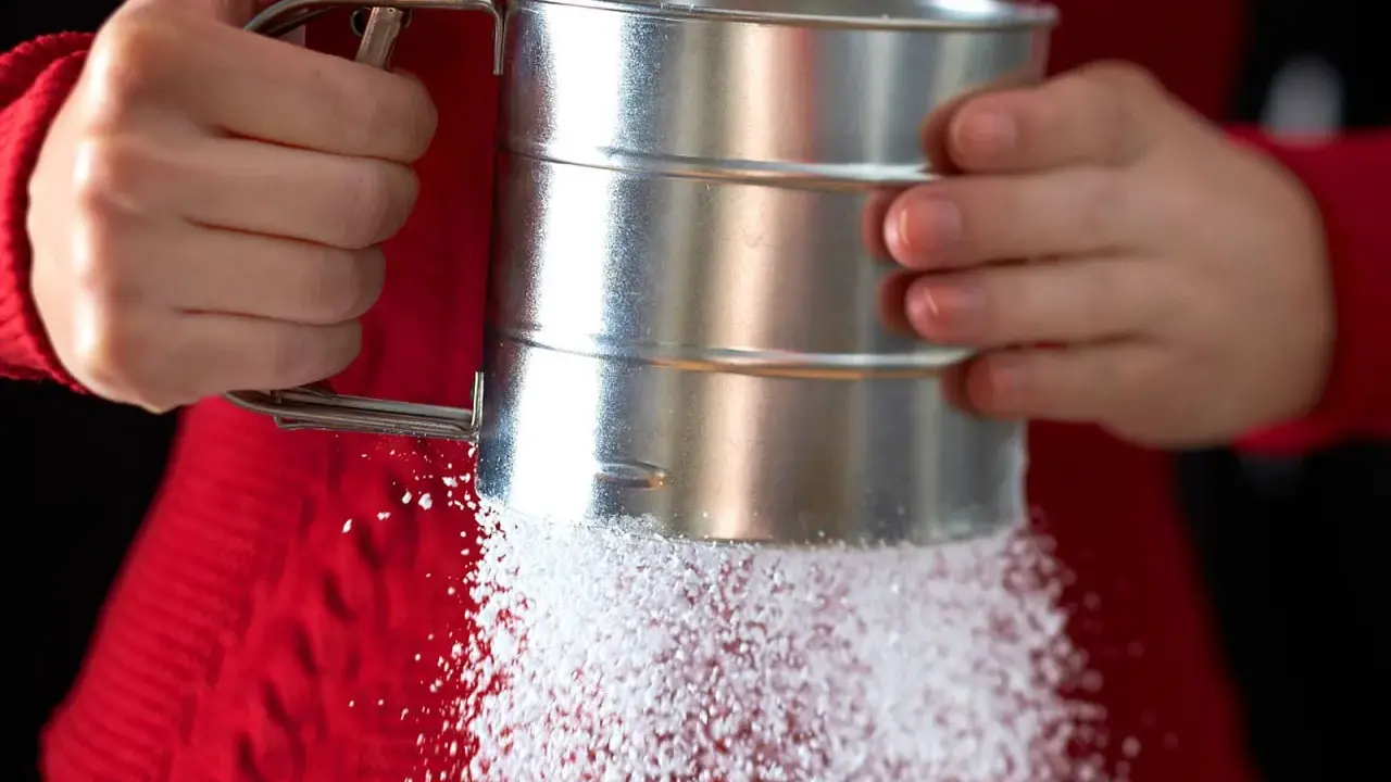 Hände halten einen Siebbecher, aus dem feiner Zucker rieselt. So einfach lässt sich puderzucker selber machen für süße Kreationen.