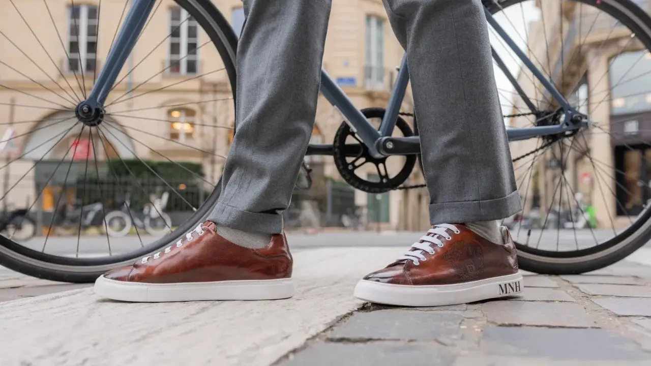 Eleganckie sneakersy do garnituru w brązowym kolorze, zestawione z szarymi spodniami, na tle miejskiego krajobrazu z rowerem.