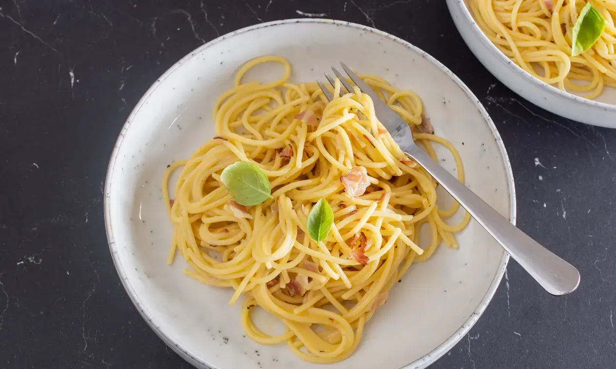 Ein Teller mit cremiger Spaghetti Carbonara, garniert mit Basilikum und Speckstückchen.