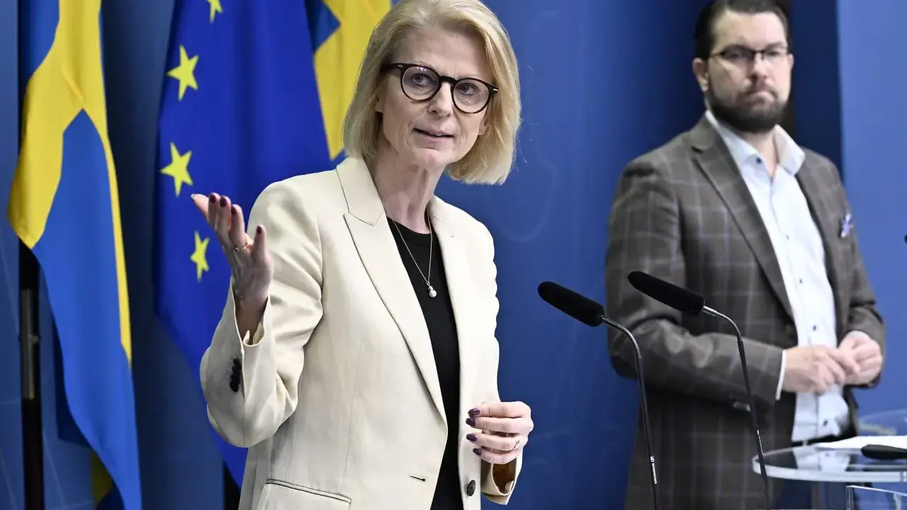 Kvinna med glasögon talar vid presskonferens med svenska och EU-flaggor bakom. Viktig information om folkräkning Sverige.