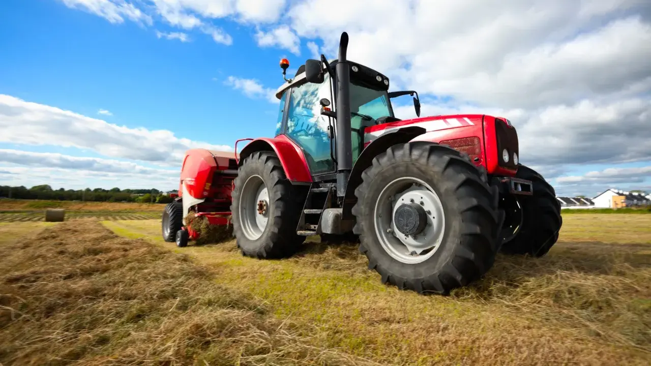 Czerwony traktor z przyczepą prasuje siano na polu. W tle błękitne niebo z chmurami. Traktorzysta w pracy.