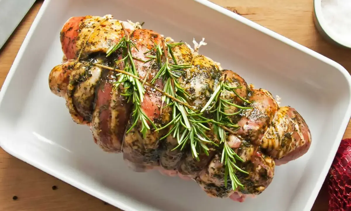 Un rôti de porc cru, assaisonné et garni de romarin frais, prêt à être cuit dans un plat blanc.