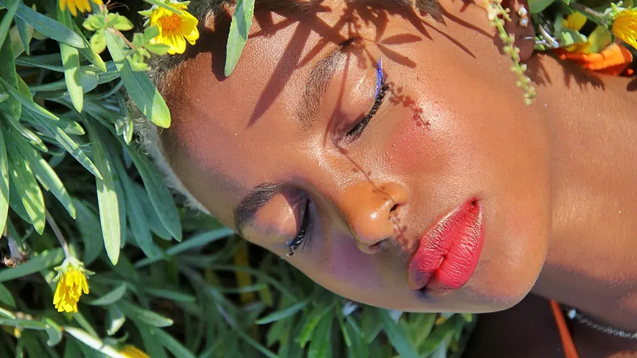Un volto immerso tra fiori, con un trucco festival audace: eyeliner blu elettrico e labbra rosse vibranti.