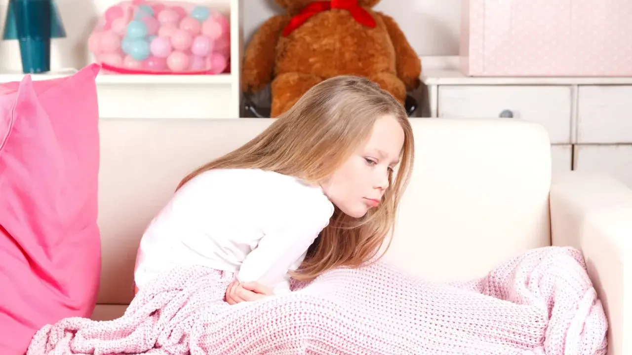 Bambina di 12 anni con mal di stomaco, rannicchiata sul divano sotto una coperta rosa.