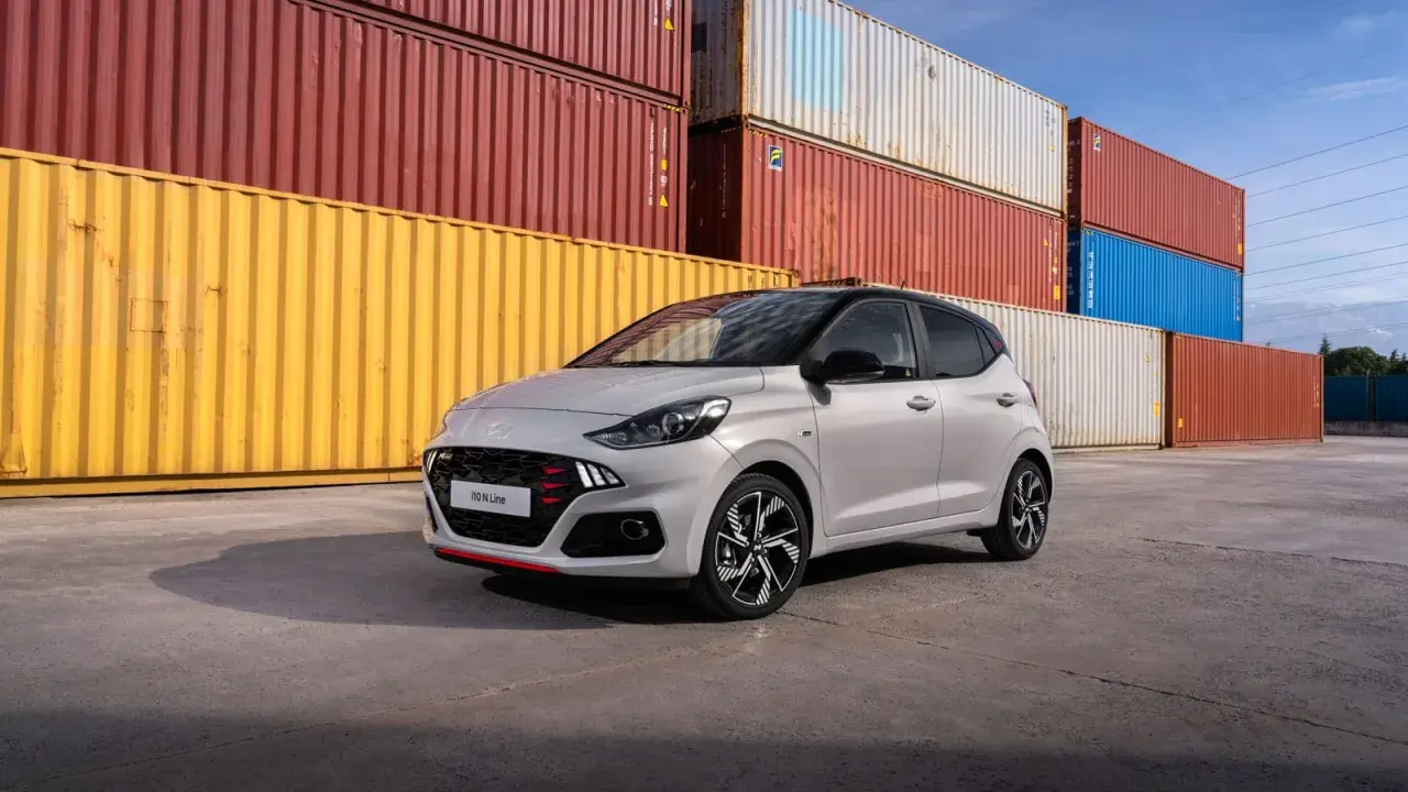 Szary Hyundai i10 N Line z czerwonymi akcentami na grillu i zderzaku, zaparkowany przed stosem kolorowych kontenerów. Sprawdź atrakcyjną hyundai i10 cena!