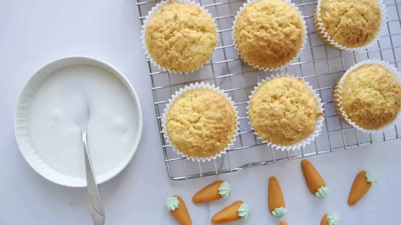 Goldbraune Karottenmuffins auf einem Gitter, daneben eine Schüssel mit Zuckerguss und süße Marzipankarotten. Perfekt für ein Karottenmuffins Rezept!