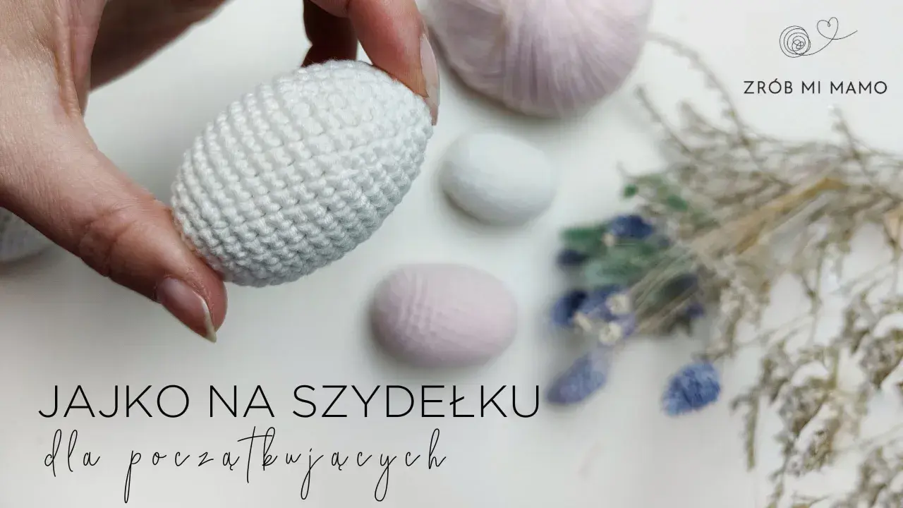 Ręcznie robione, białe jajko na szydełku, idealne na początek przygody z szydełkowaniem. W tle pastelowe włóczki i suszone kwiaty.