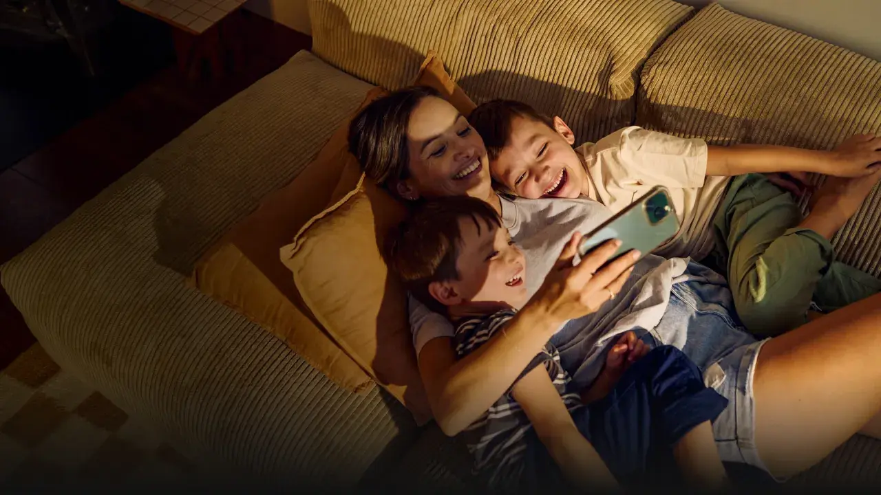 A mother and two sons laugh while looking at a smartphone on a couch.