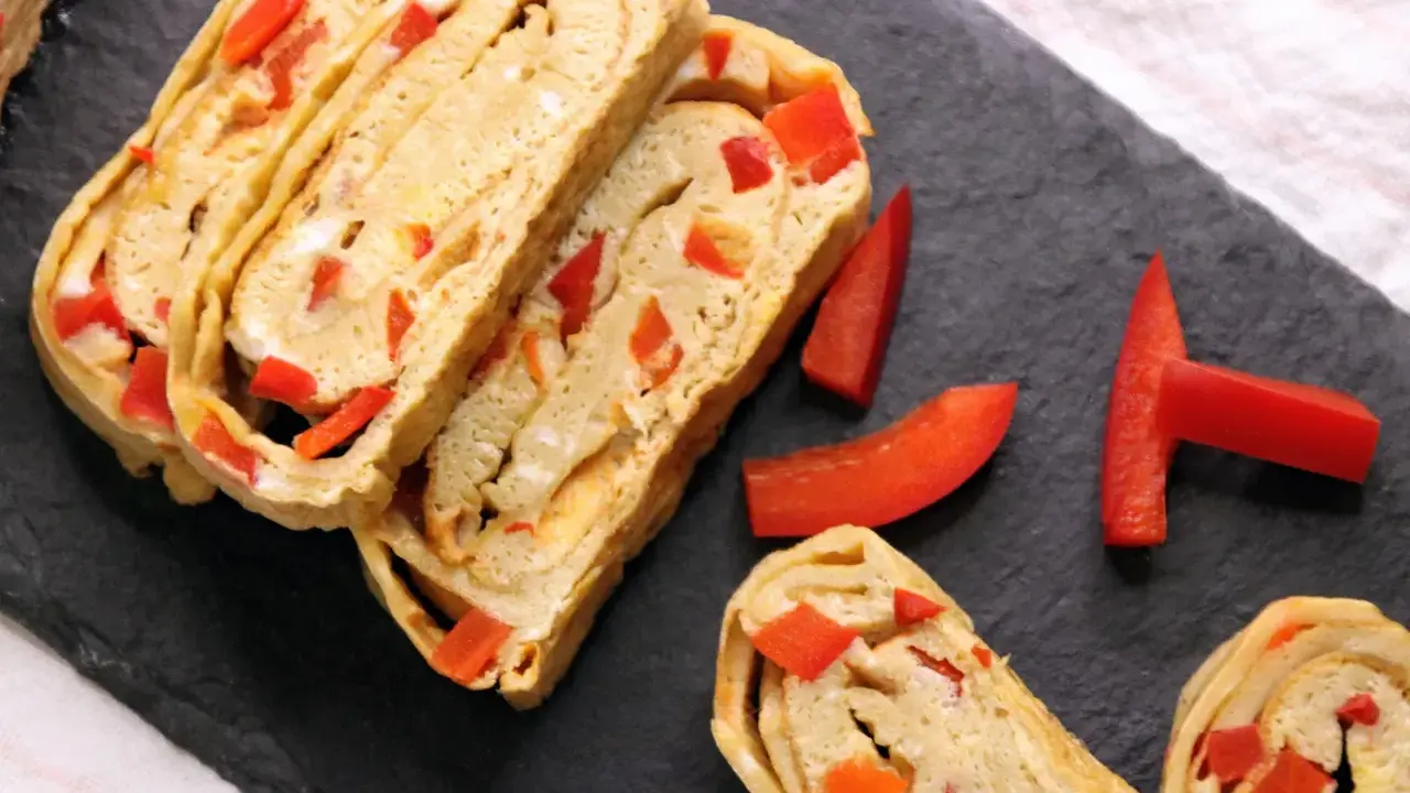Roladki omletu tamago z kawałkami czerwonej papryki, ułożone na czarnej desce.