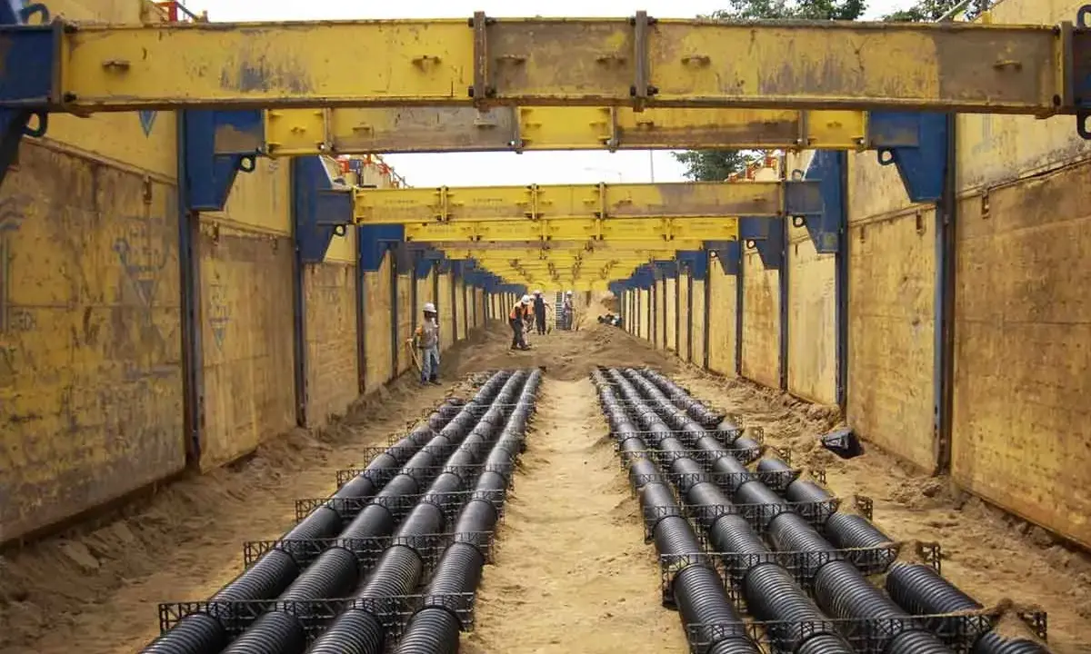 Trabajadores instalan tuberías en una zanja profunda, con estructuras metálicas amarillas para entibar zanjas y prevenir derrumbes.