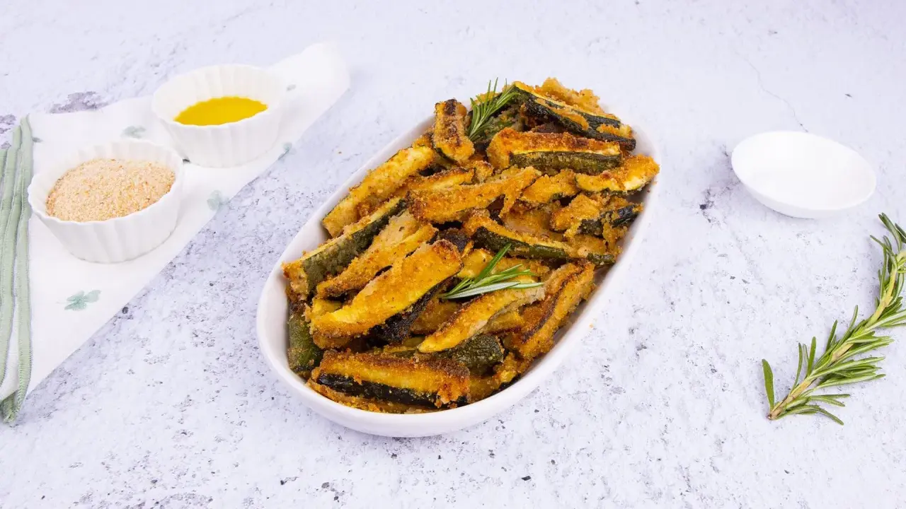 Zucchine gratinate al forno, dorate e croccanti, servite in un piatto ovale con rosmarino fresco. Accanto, ciotoline con pangrattato e olio.