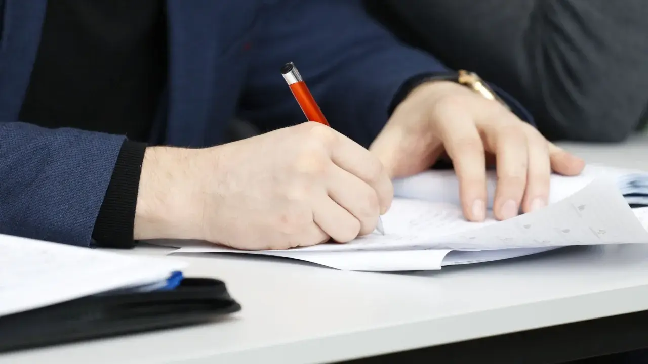 Ręka pisze długopisem po papierze, przygotowując przykładowy artykuł po angielsku na maturę.