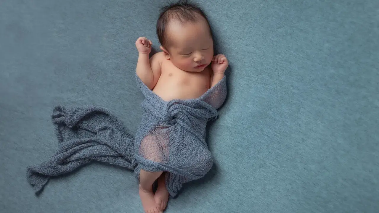 Neonato dorme avvolto in un telo blu, con le braccia sollevate e i pugni chiusi.