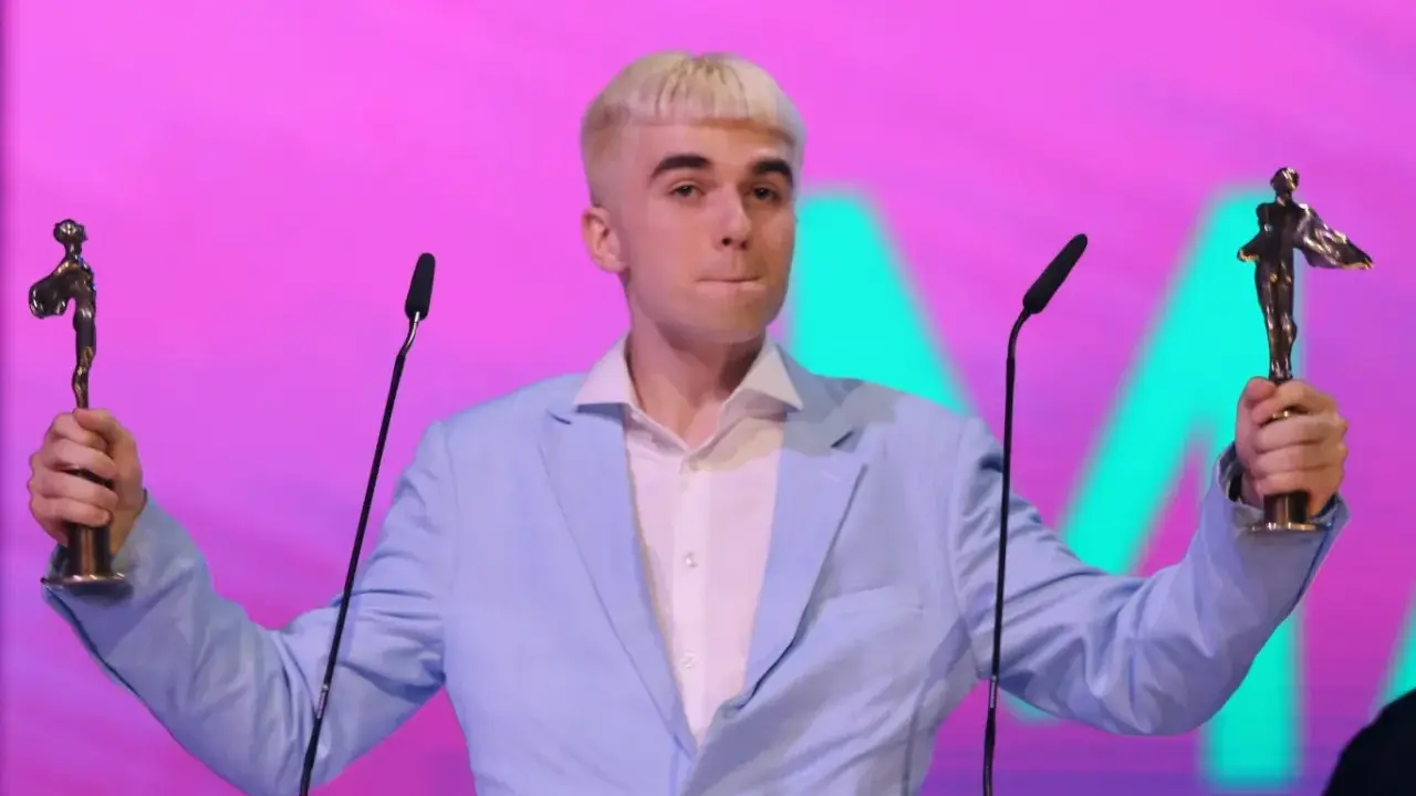 Mc Mata z dwoma statuetkami na scenie, w jasnoniebieskiej marynarce i białej koszuli.