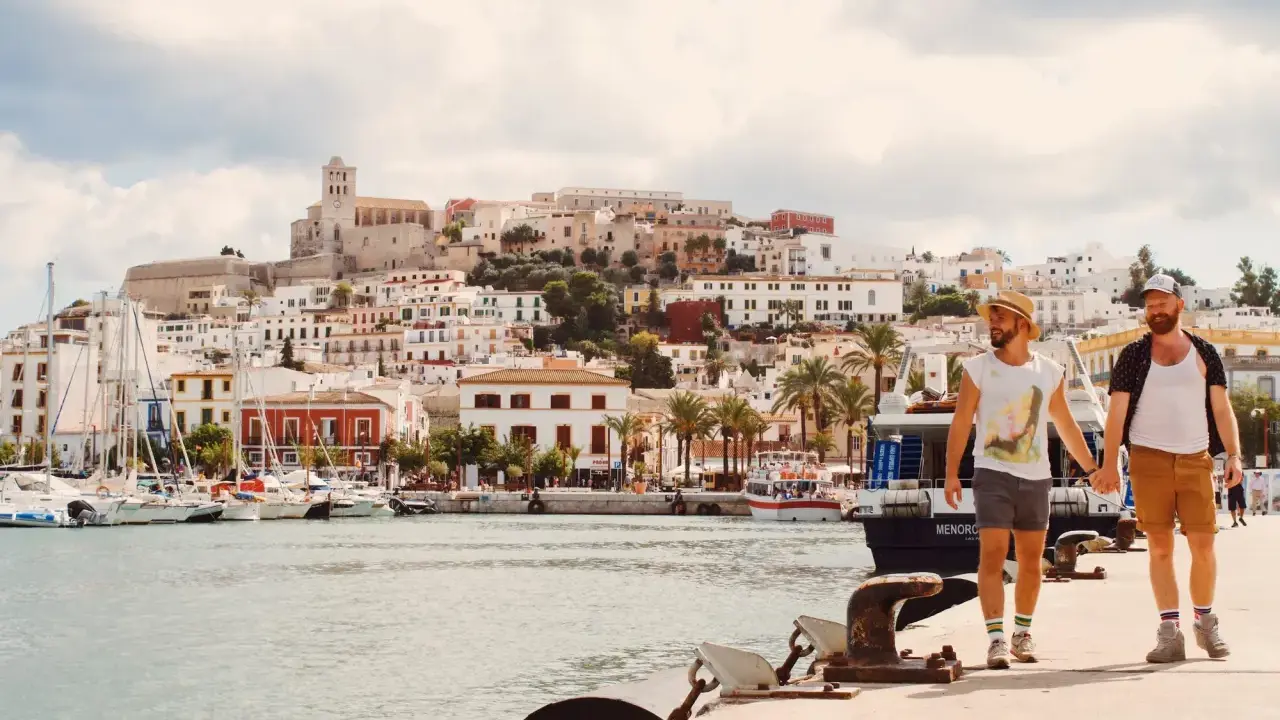 A gay couple walks hand-in-hand along a harbor in Spain, with a white-washed town and ancient fortress rising behind them.