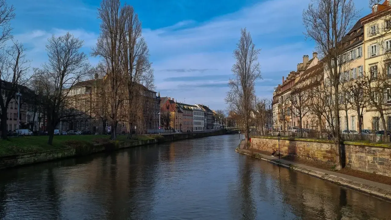 Spokojna rzeka w **Strasburgu** płynie obok kamiennych nabrzeży i kolorowych kamienic. Drzewa bez liści tworzą zimowy krajobraz.
